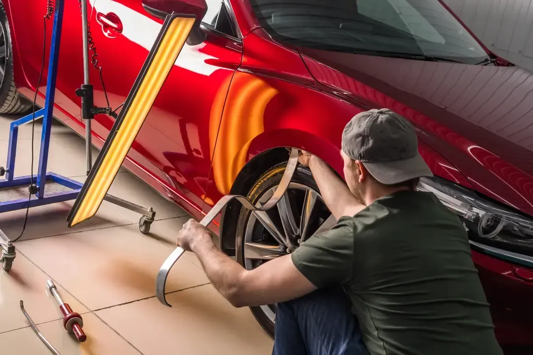 A technician uses a specialized metal tool to perform paintless dent repair on the front fender of a red car.