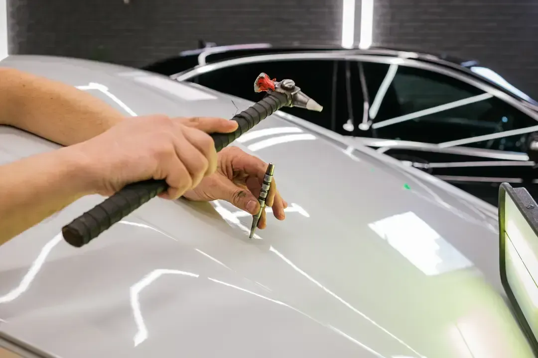 A technician uses a specialized tapping tool to perform paintless dent repair on a silver car roof in a workshop.