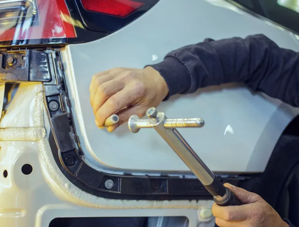 A mechanic holds a metal stud welding tool near the rear fender of a white car during auto body repair.