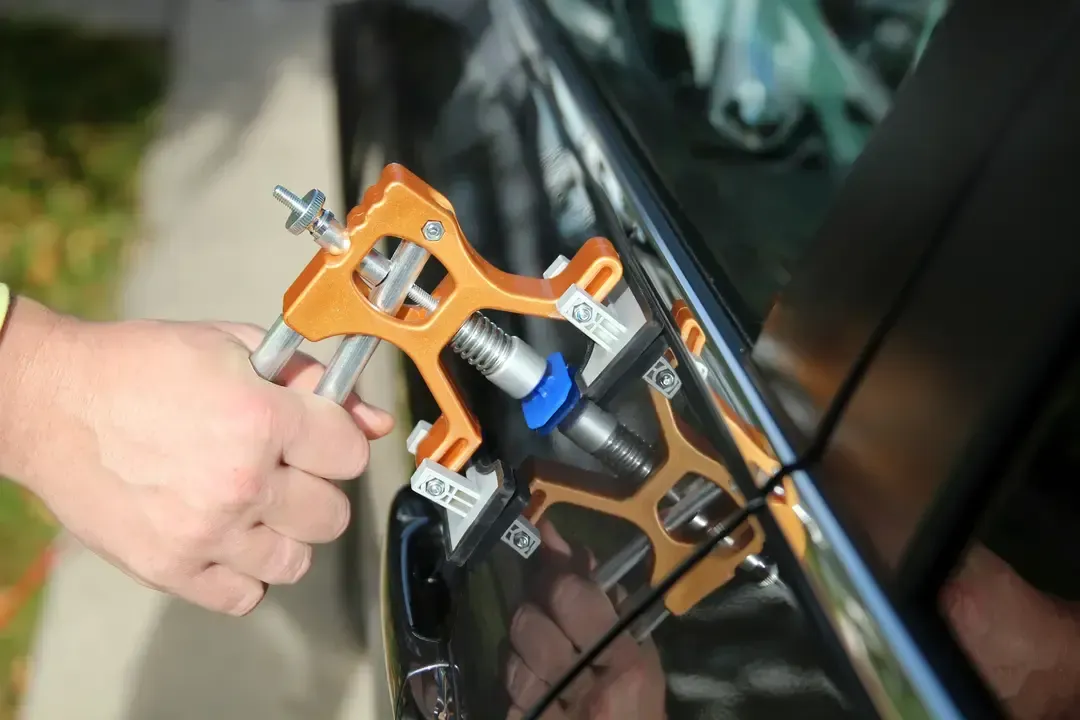A person uses a gold-colored dent removal tool on the door panel of a black car.