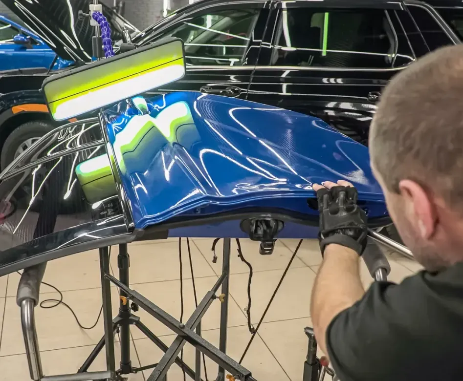 A technician uses a light board to perform paintless dent repair on a detached blue car hood in a workshop.