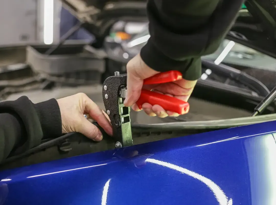 A person uses a red-handled professional dent puller tool to repair a small dent on a blue car hood.