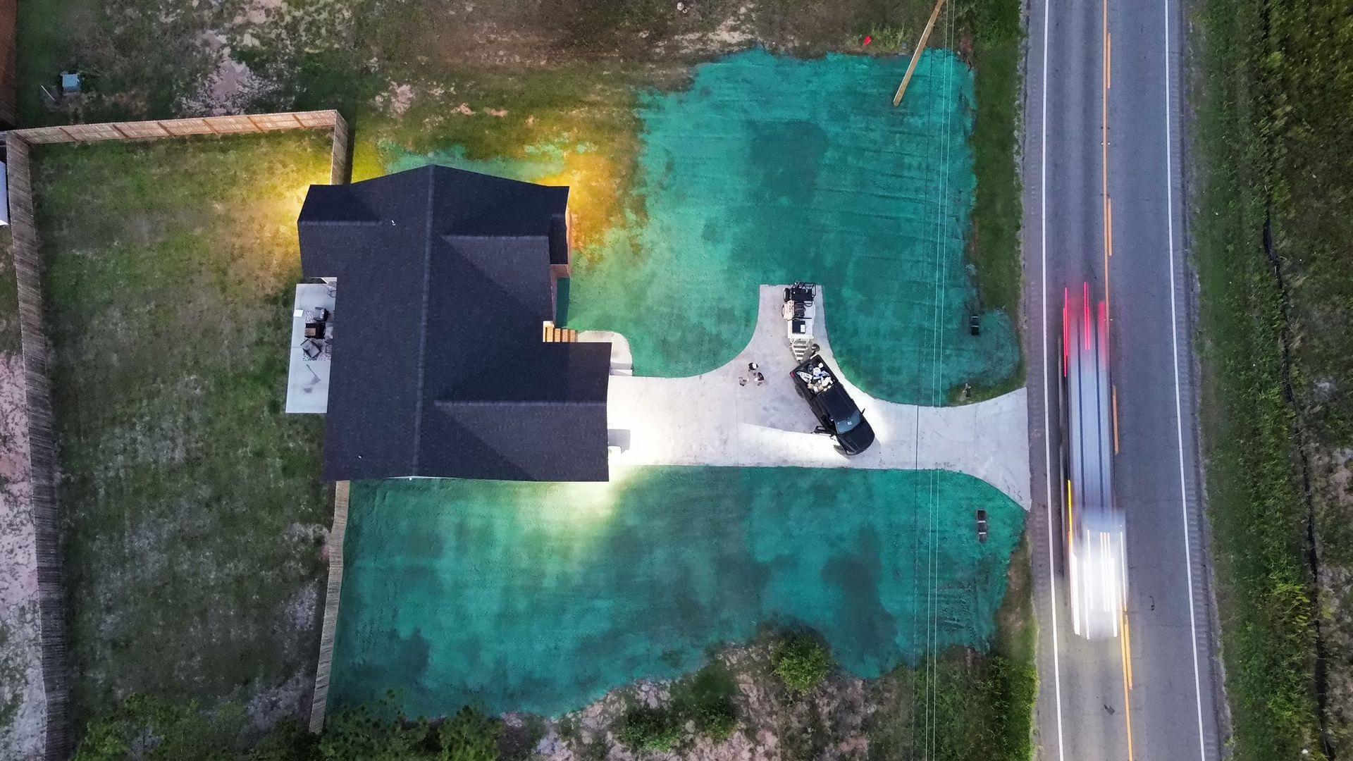 Aerial view of a house with a dark roof, green lawn, driveway, and road beside it.
