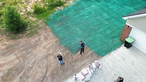 Two people standing on a dirt lot beside a house, with green netting and stacked bags nearby.