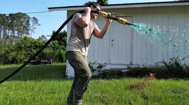 Person carrying a hose over a shoulder sprays water beside a white building on a sunny lawn.