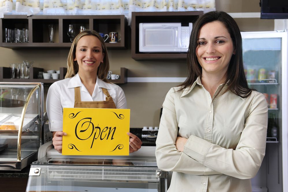 two young women stand by their open business sign