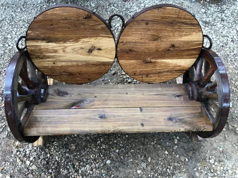 A wooden bench made out of wagon wheels is sitting on a gravel road.