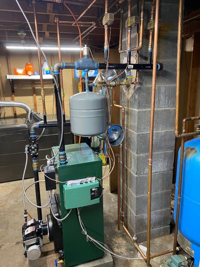 A green residential boiler system with connected copper pipes, a gray expansion tank, and a blue water tank in a basement.
