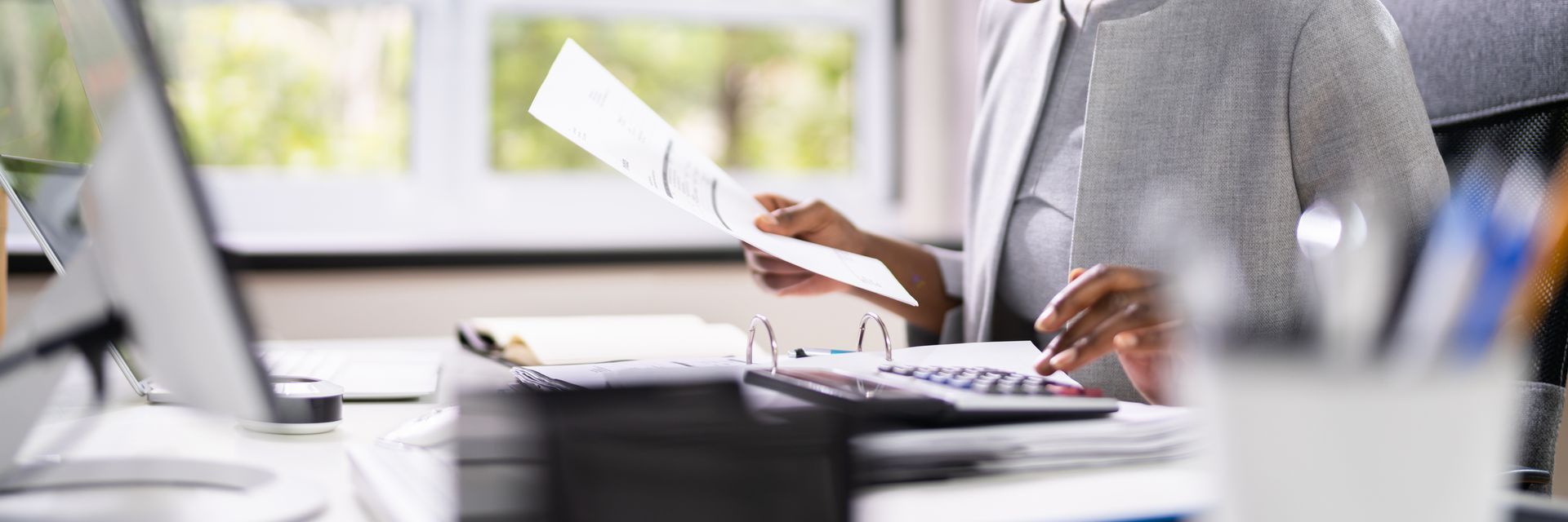 A woman is sitting at a desk in front of a computer holding a piece of paper.