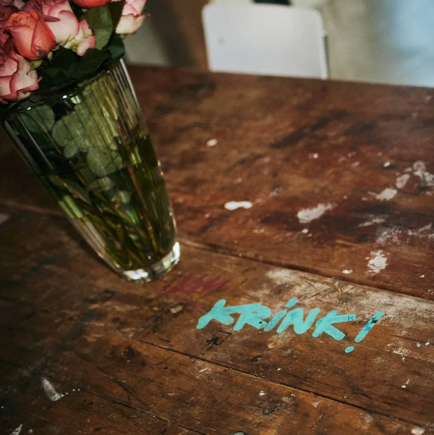 Eine Vase mit Rosen auf einem abgenutzten Holztisch. Türkisfarbenes Graffiti mit der Aufschrift „krink!“.