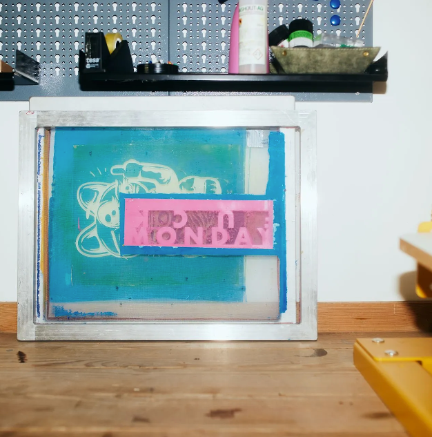 Ein Siebdruckrahmen mit blauer und rosa Farbe zeigt ein Motiv mit der Aufschrift „ICH HASSE MONTAG“. 