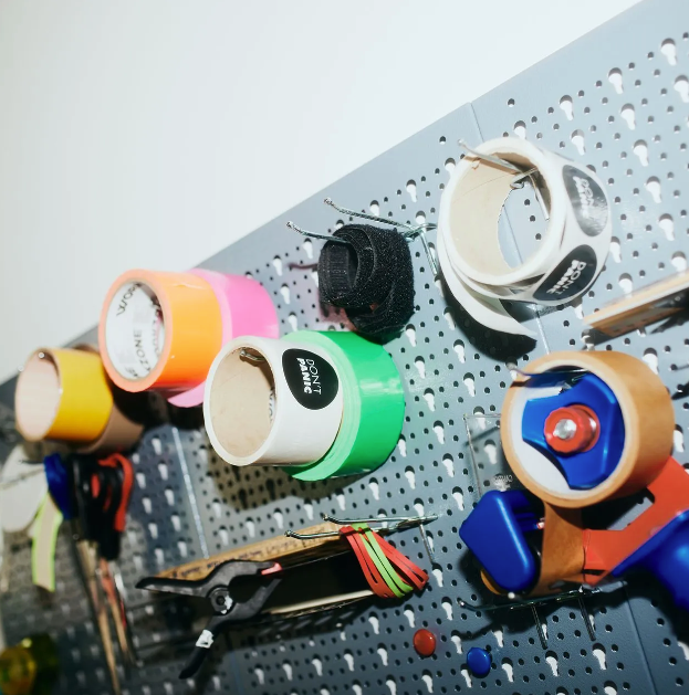 Lochwand mit Klebebandrollen, Etiketten und Werkzeugen. Verschiedene Farben.