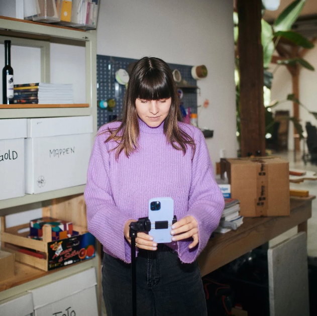 Frau in lila Pullover filmt mit Smartphone in einem unordentlichen Arbeitsbereich.