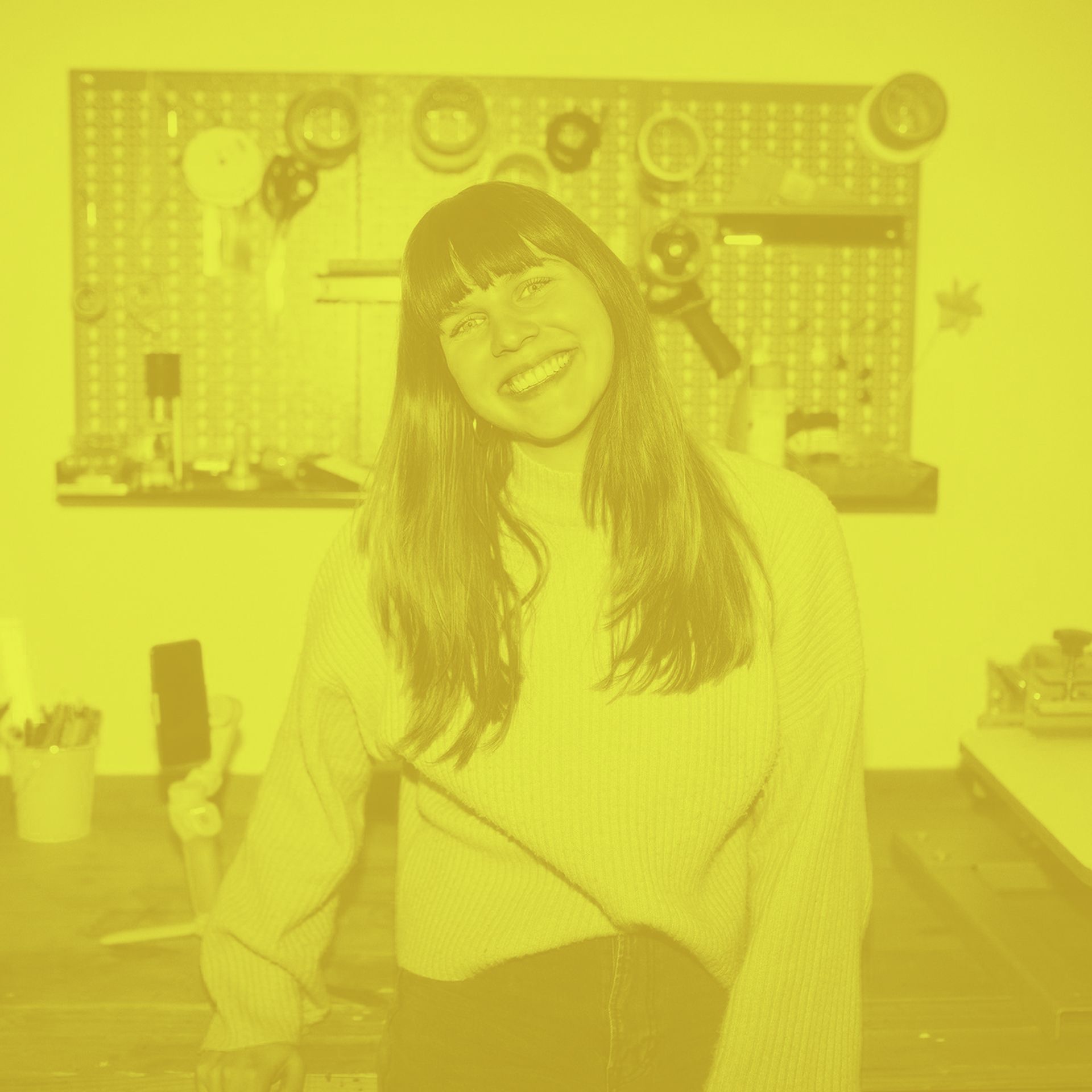 Eine Frau lächelt, an einen Tisch in einer Werkstatt gelehnt, umgeben von Werkzeugen und Material.