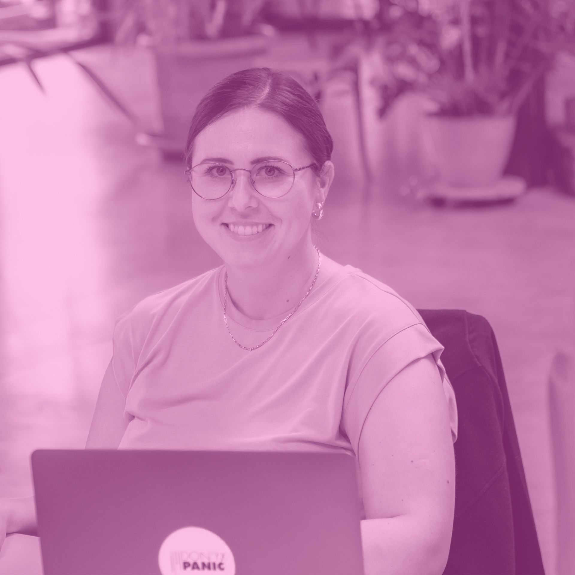 Frau lächelt in die Kamera, trägt eine Brille und sitzt an einem Schreibtisch mit einem Laptop. Rosa Farbüberlagerung.