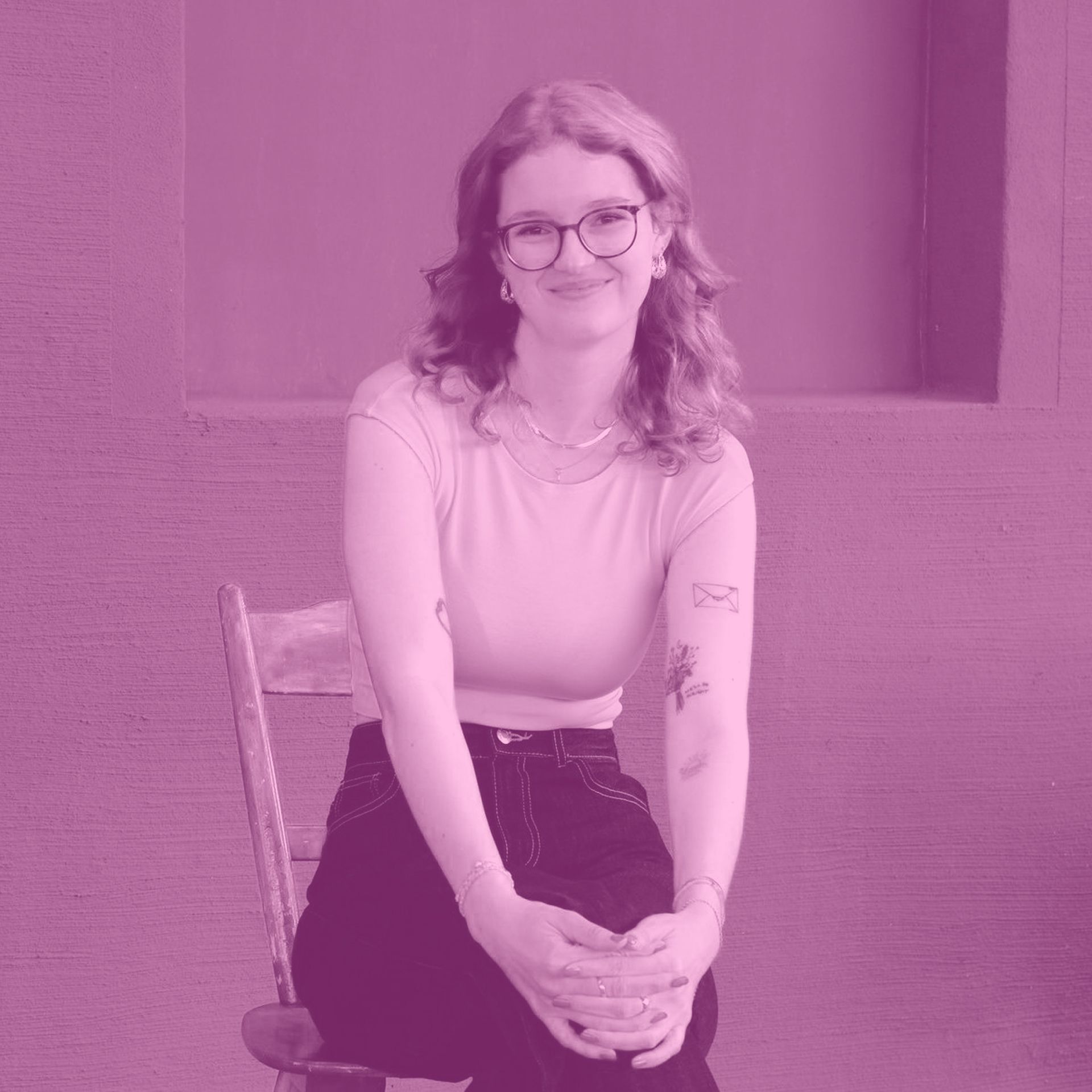 Frau mit Brille und Tätowierungen sitzt lächelnd auf einem Stuhl. Rosa Farbton.