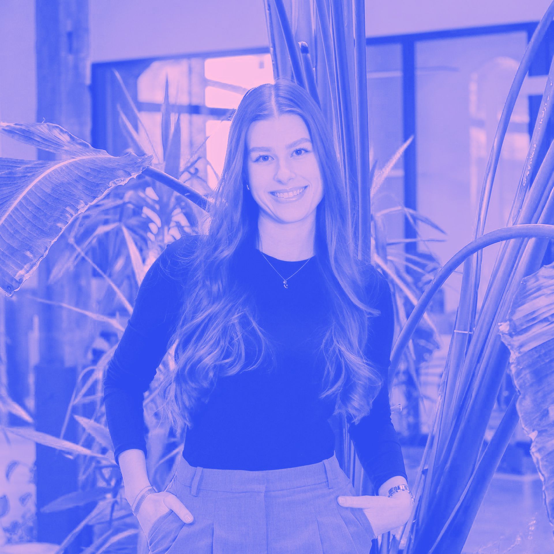 Frau mit langen braunen Haaren, lächelnd, trägt ein schwarzes Hemd und eine graue Hose und steht im Freien.