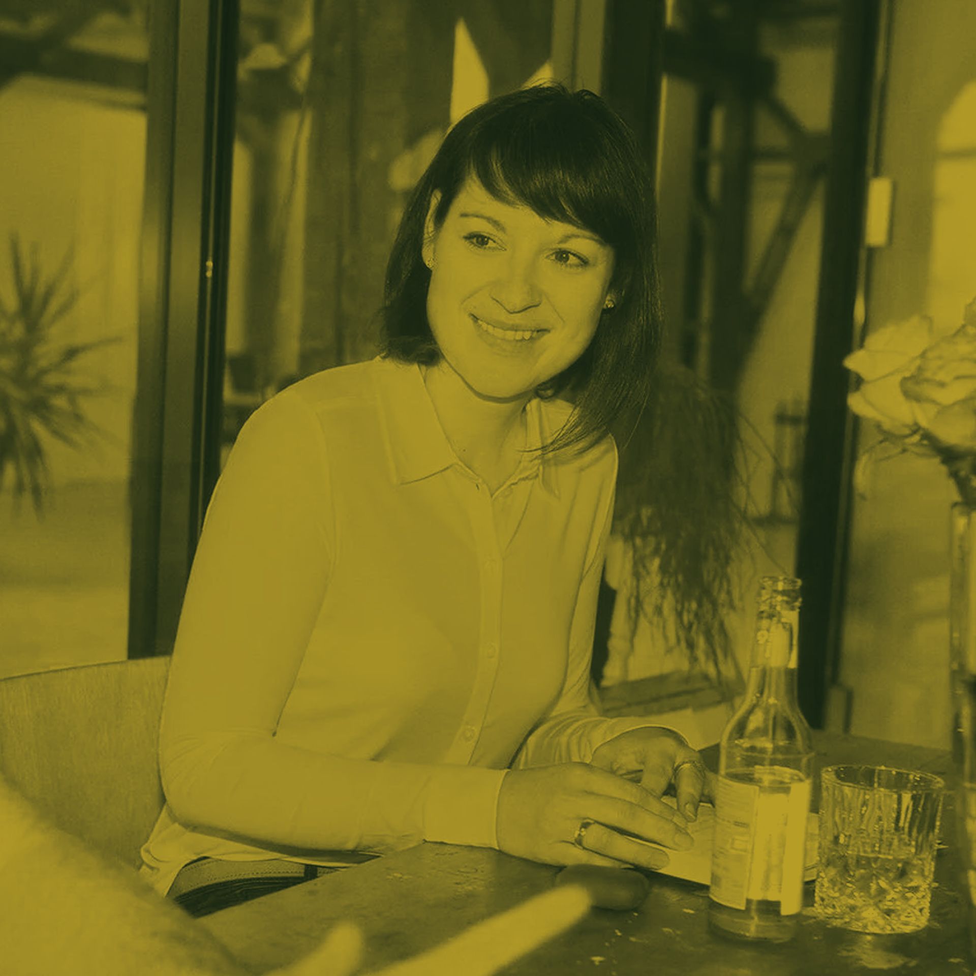 Eine Frau sitzt lächelnd an einem Tisch; vor ihr stehen eine Flasche und ein Glas. Innenaufnahme, gelbliches Licht.