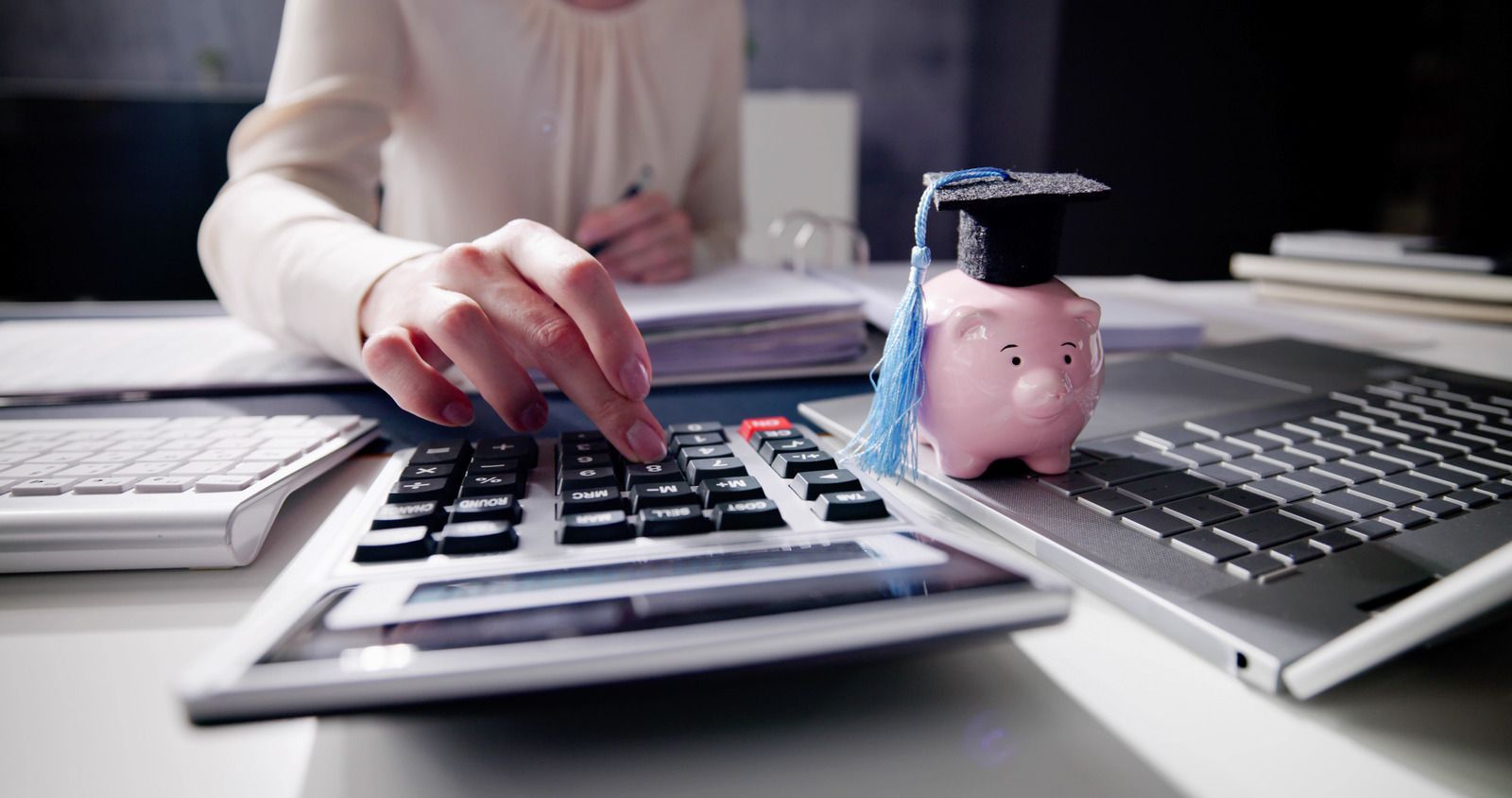 A woman is using a calculator next to a piggy bank with a graduation cap on it.