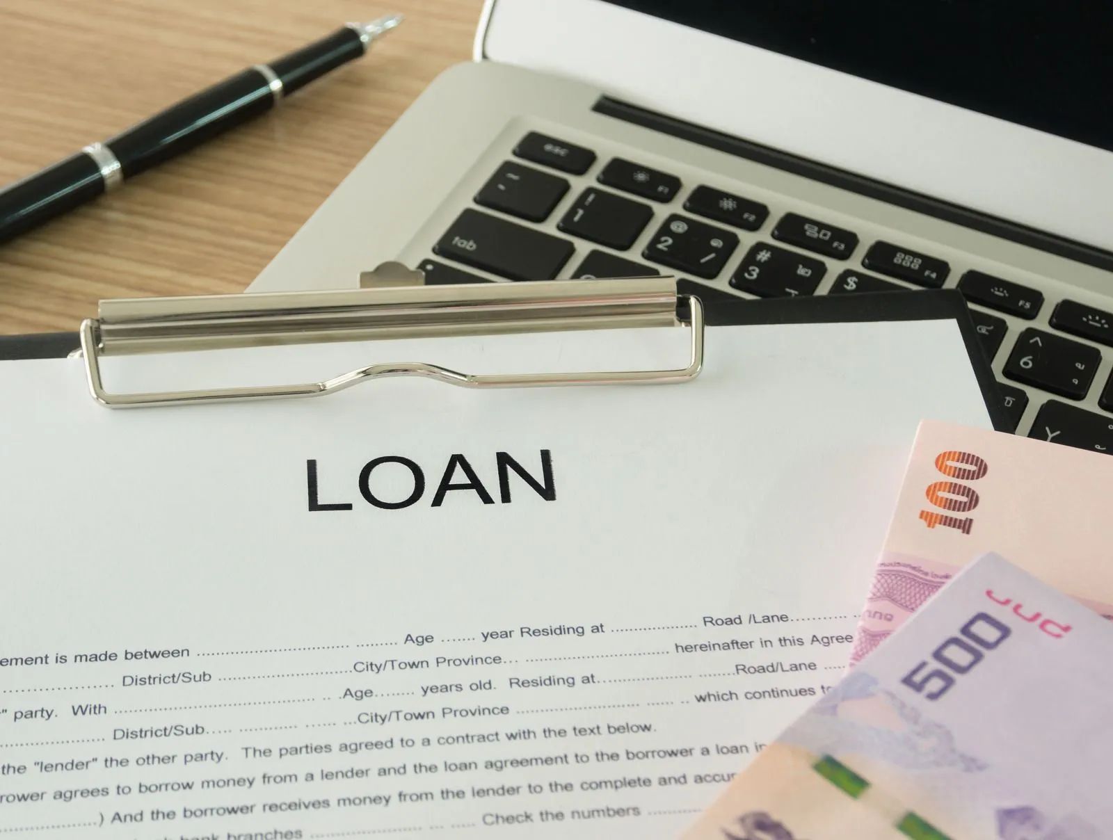 Loan document on clipboard, laptop, pen, and cash on a wooden desk.