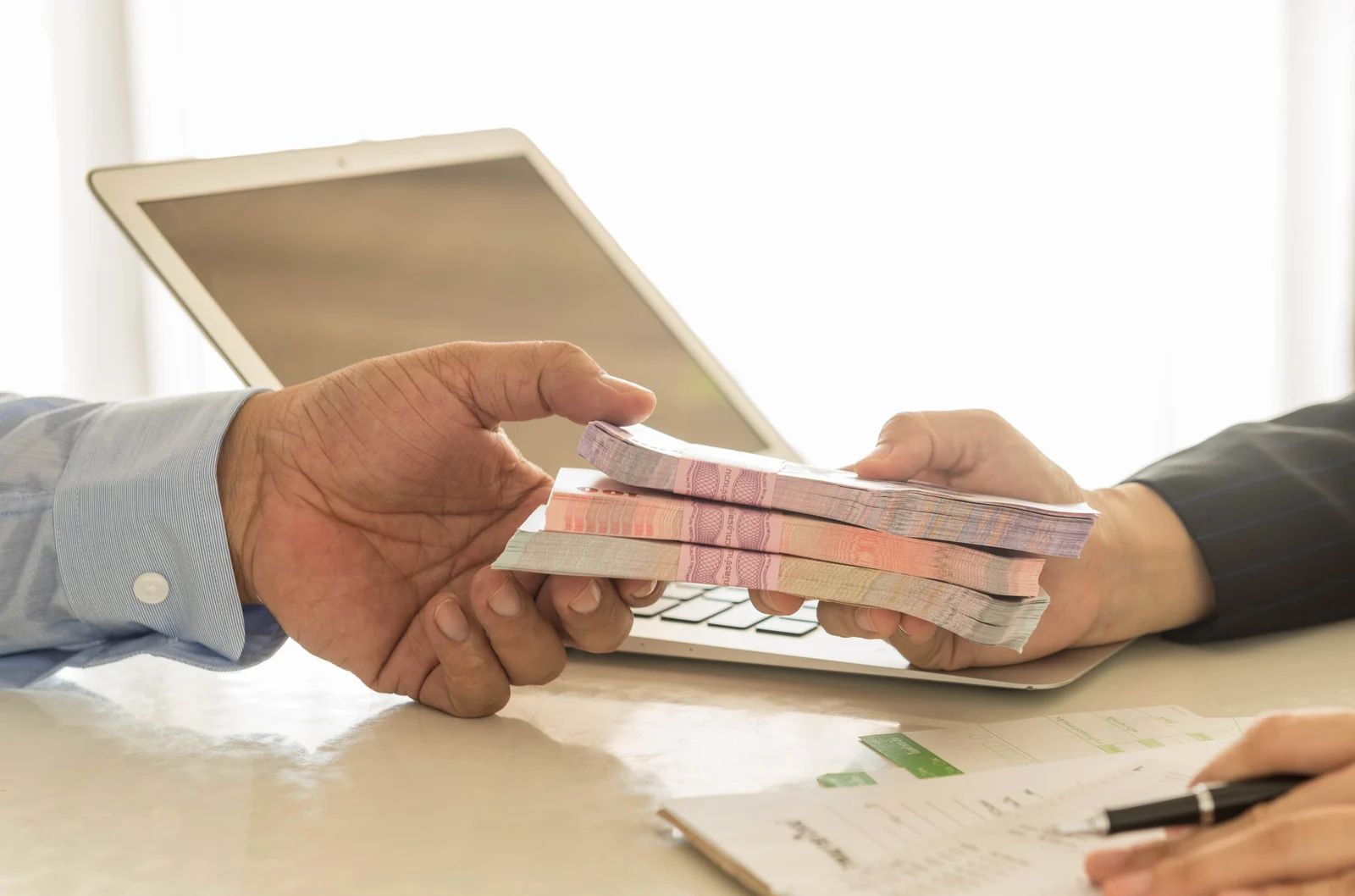Hands exchanging stack of banknotes, laptop and documents on table.