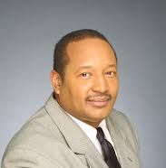 Man in light grey suit and black tie, smiling. Studio setting with grey background.
