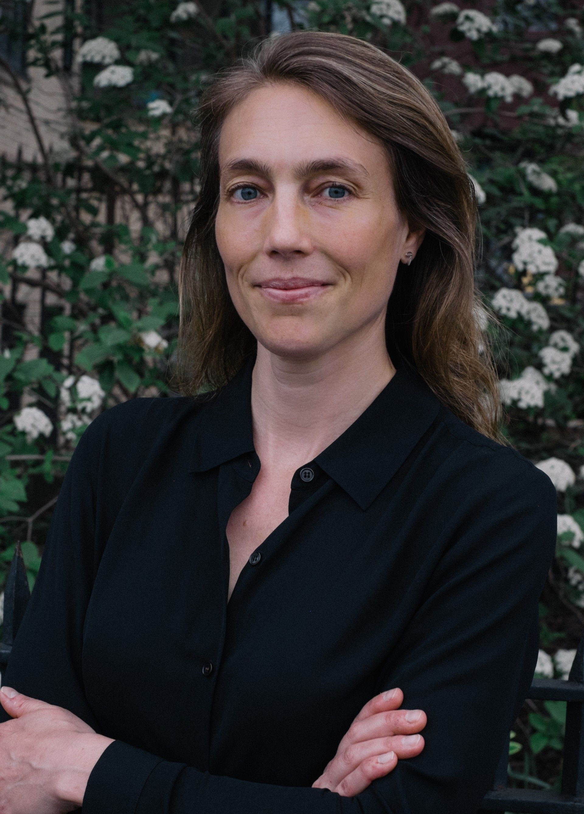 Woman with long brown hair, arms crossed, wearing a black shirt, standing outside with white flowers in background.