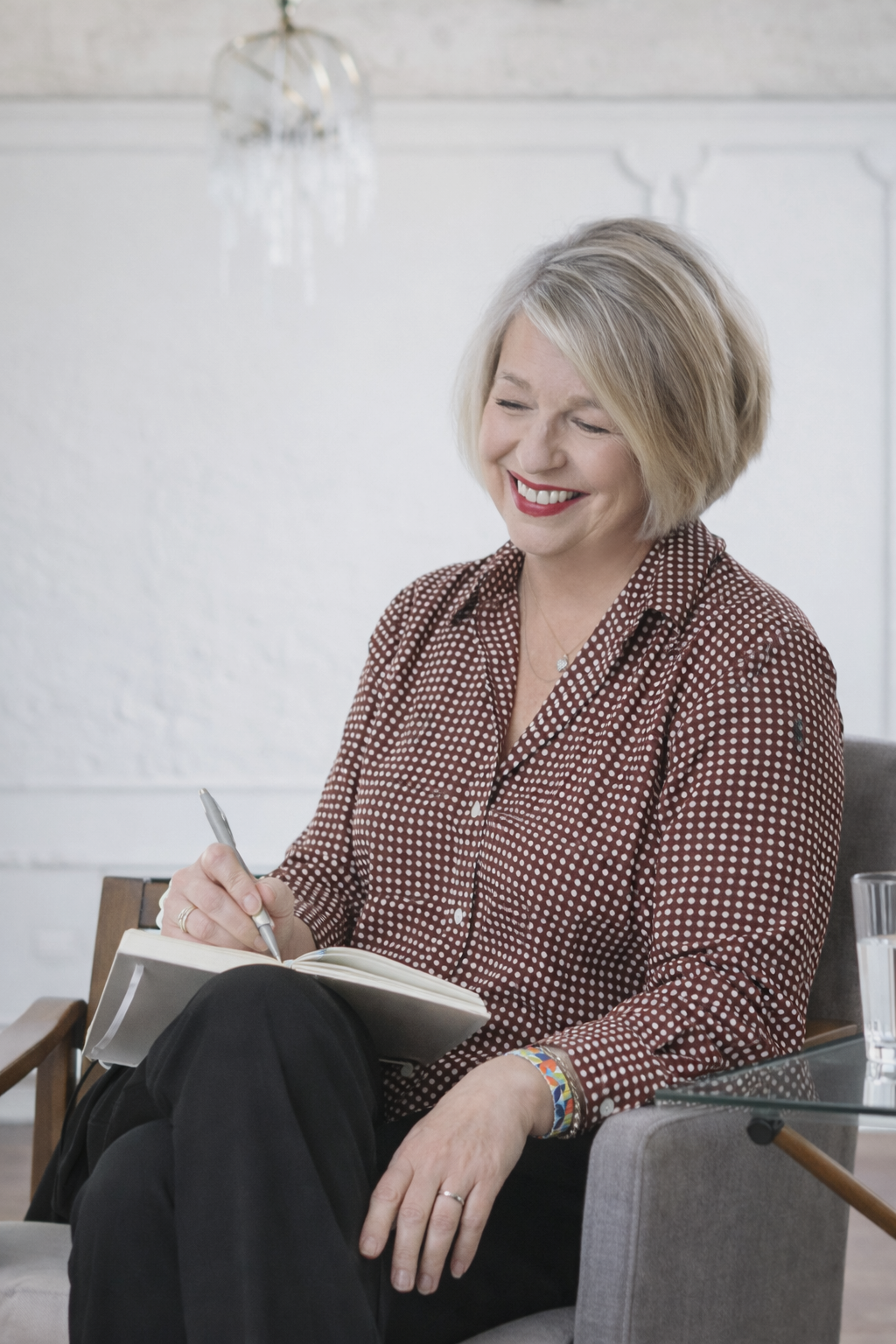 Woman writing in a notebook, smiling. Wearing patterned shirt, sitting in armchair, bright room.