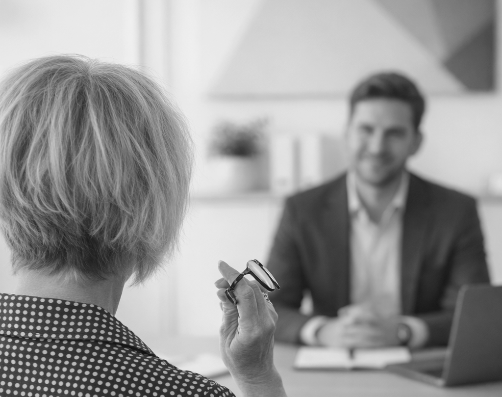 Woman in patterned jacket holds glasses, facing a blurred man in a suit, seated at a desk.