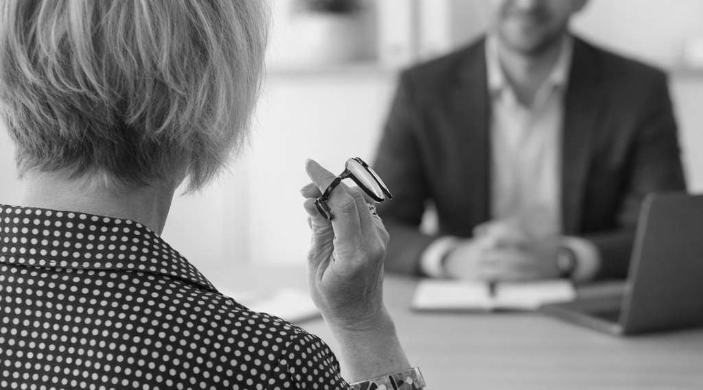 Woman in patterned top, holding glasses, facing a man in a suit at a desk; office setting.
