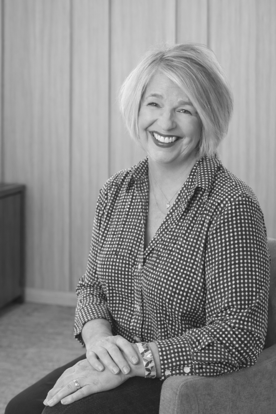 Woman in patterned shirt smiles, sitting indoors.