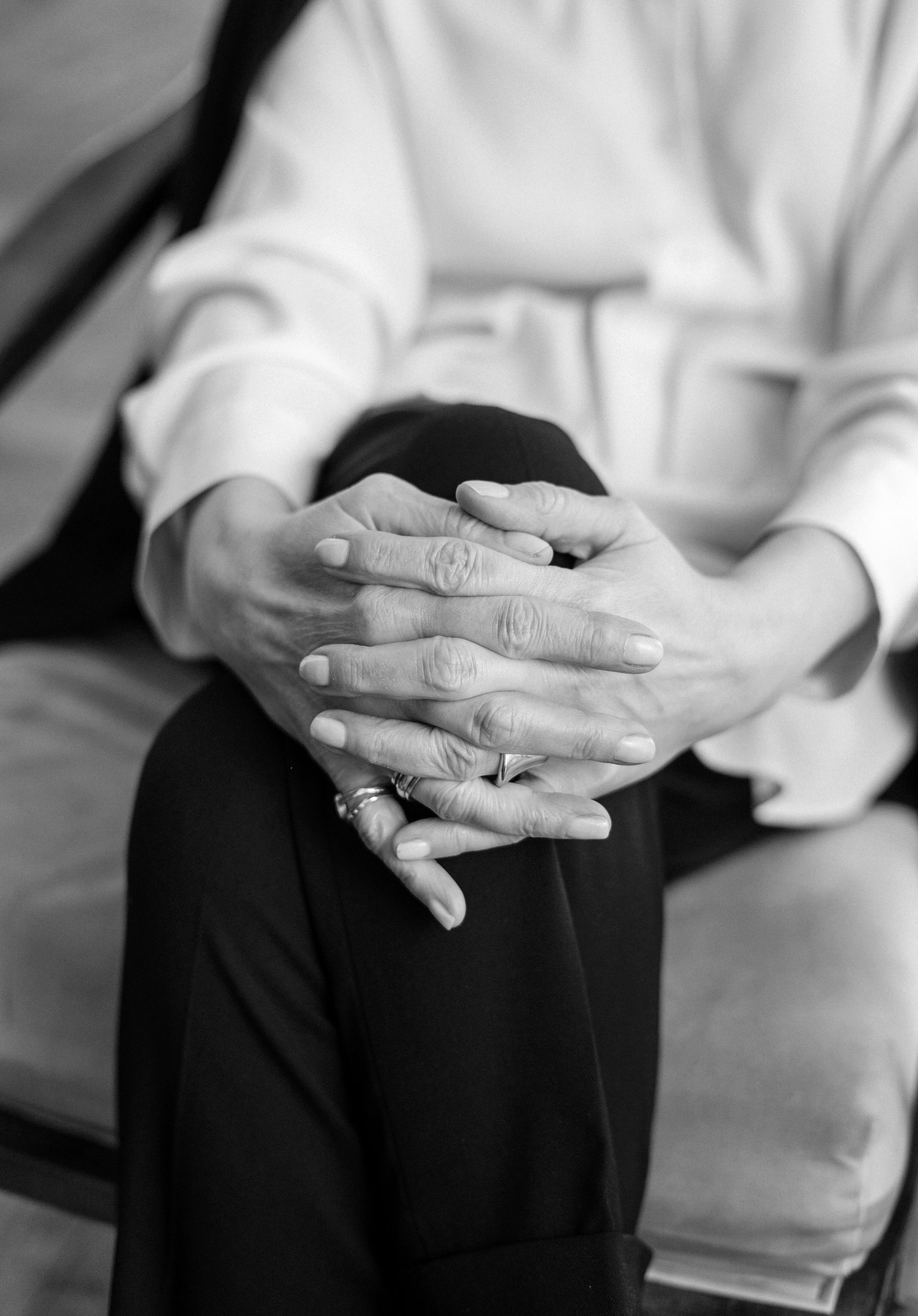 Hands clasped on lap. Person wearing white shirt and black pants, seated on a cushion.