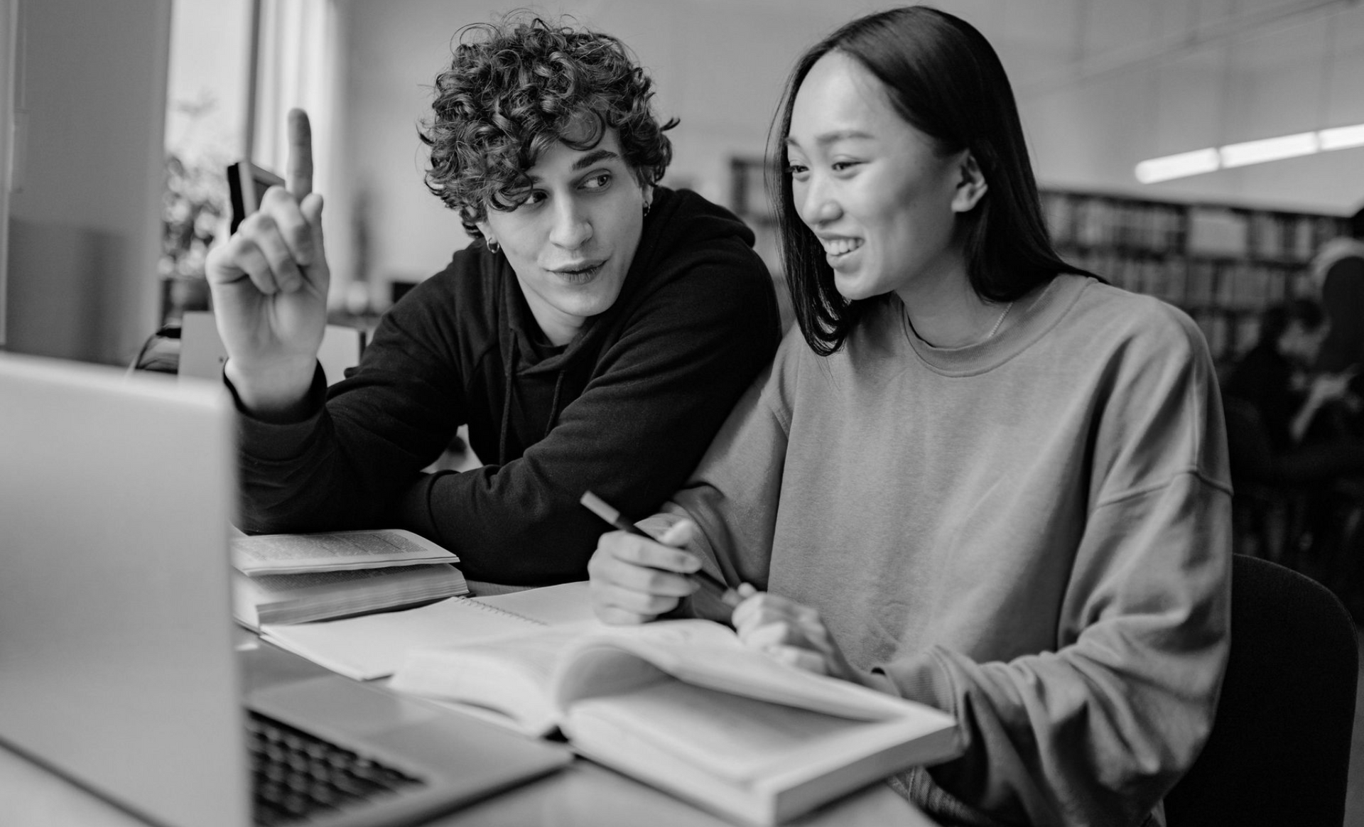 Two students studying together in a library, looking at a laptop and open book. One points upwards.