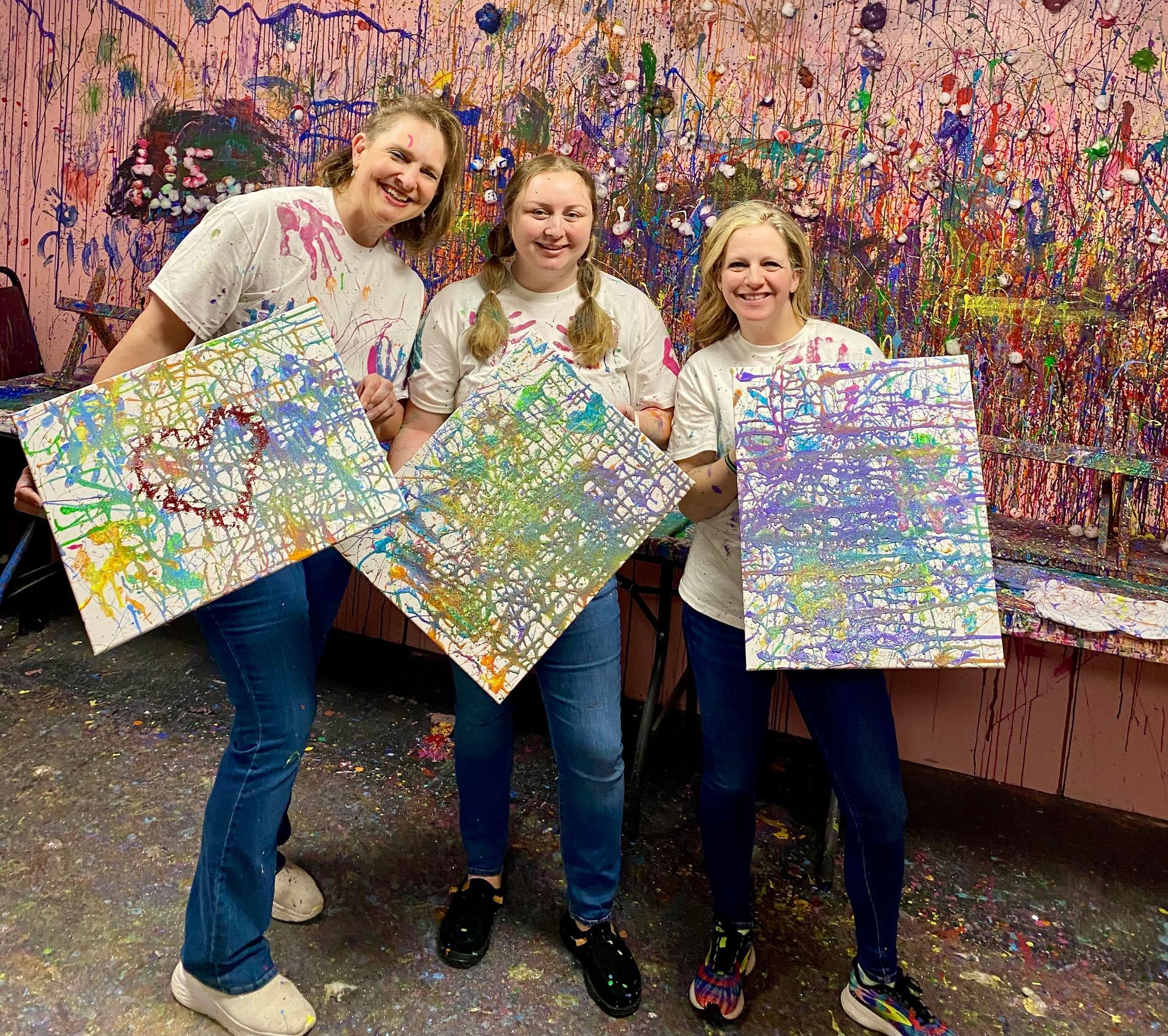 Three women are holding paintings in front of a wall covered in paint.