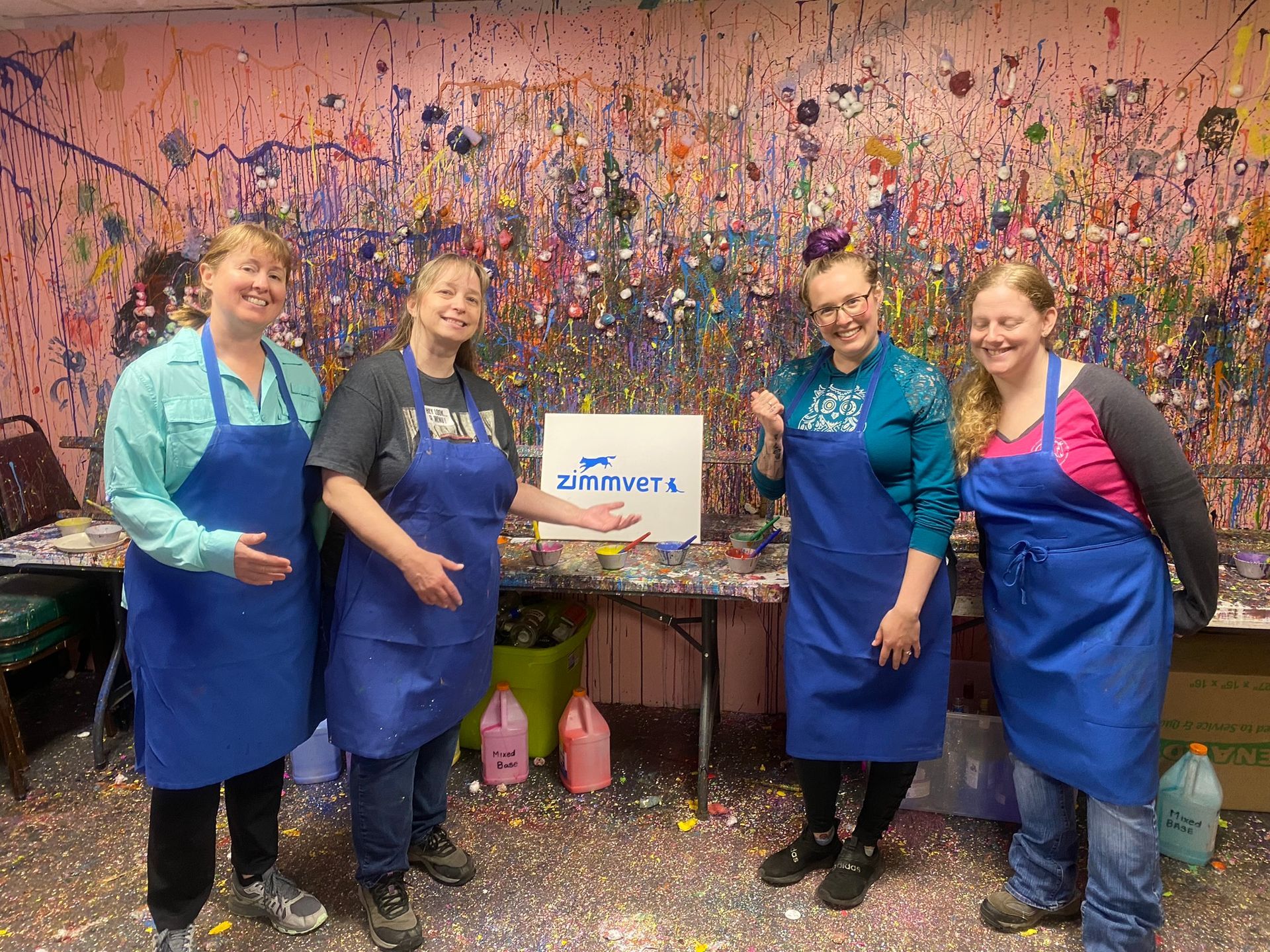 A group of women wearing blue aprons are standing in front of a wall covered in paint.