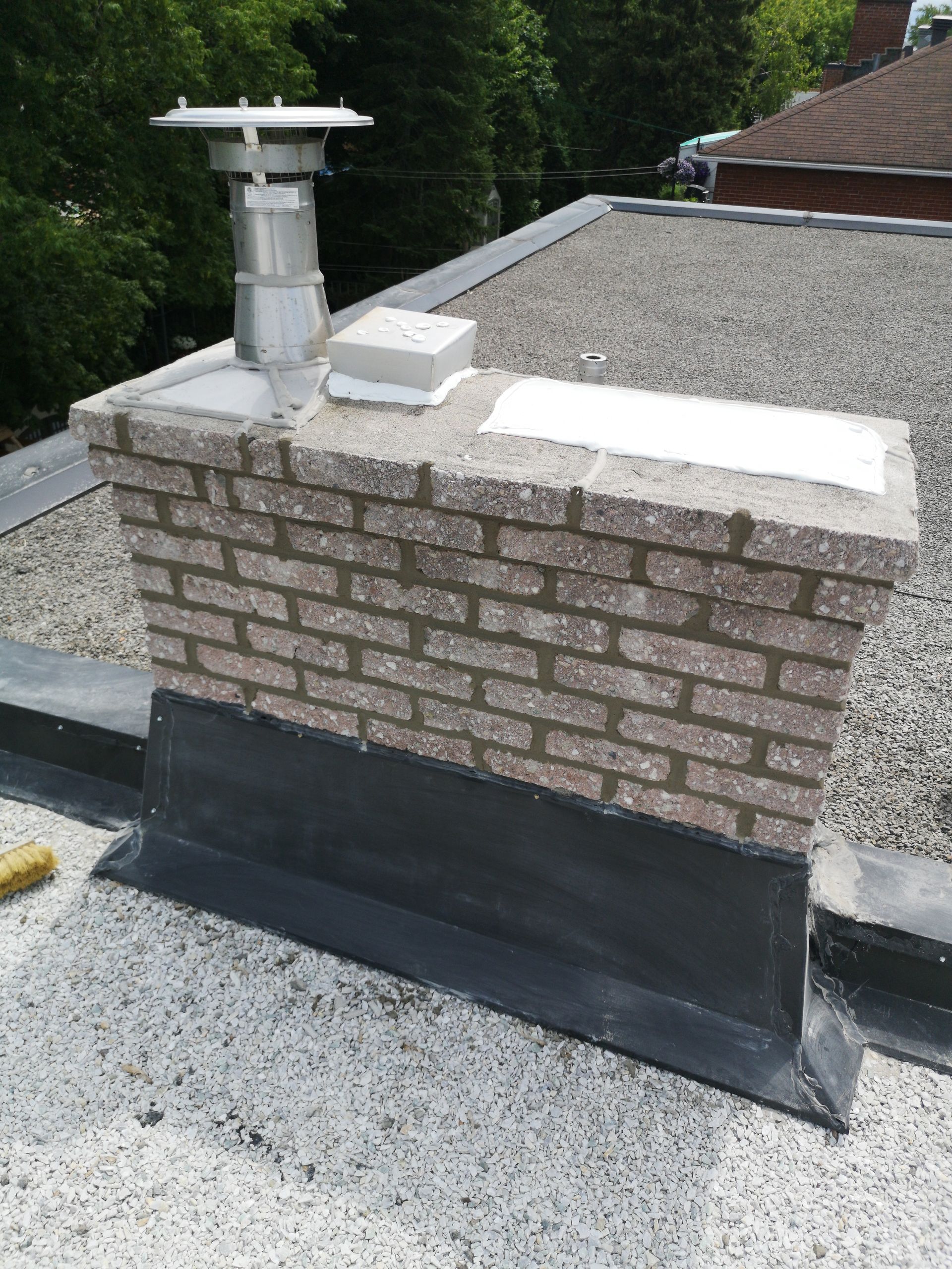 Une cheminée en briques sur un toit plat en gravier, avec un conduit de ventilation métallique et des plaques d'étanchéité
