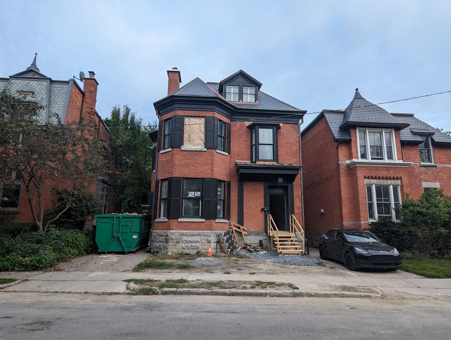 Maison en briques rouges en construction; fenêtres barricadées; toit noir; benne à ordures verte.