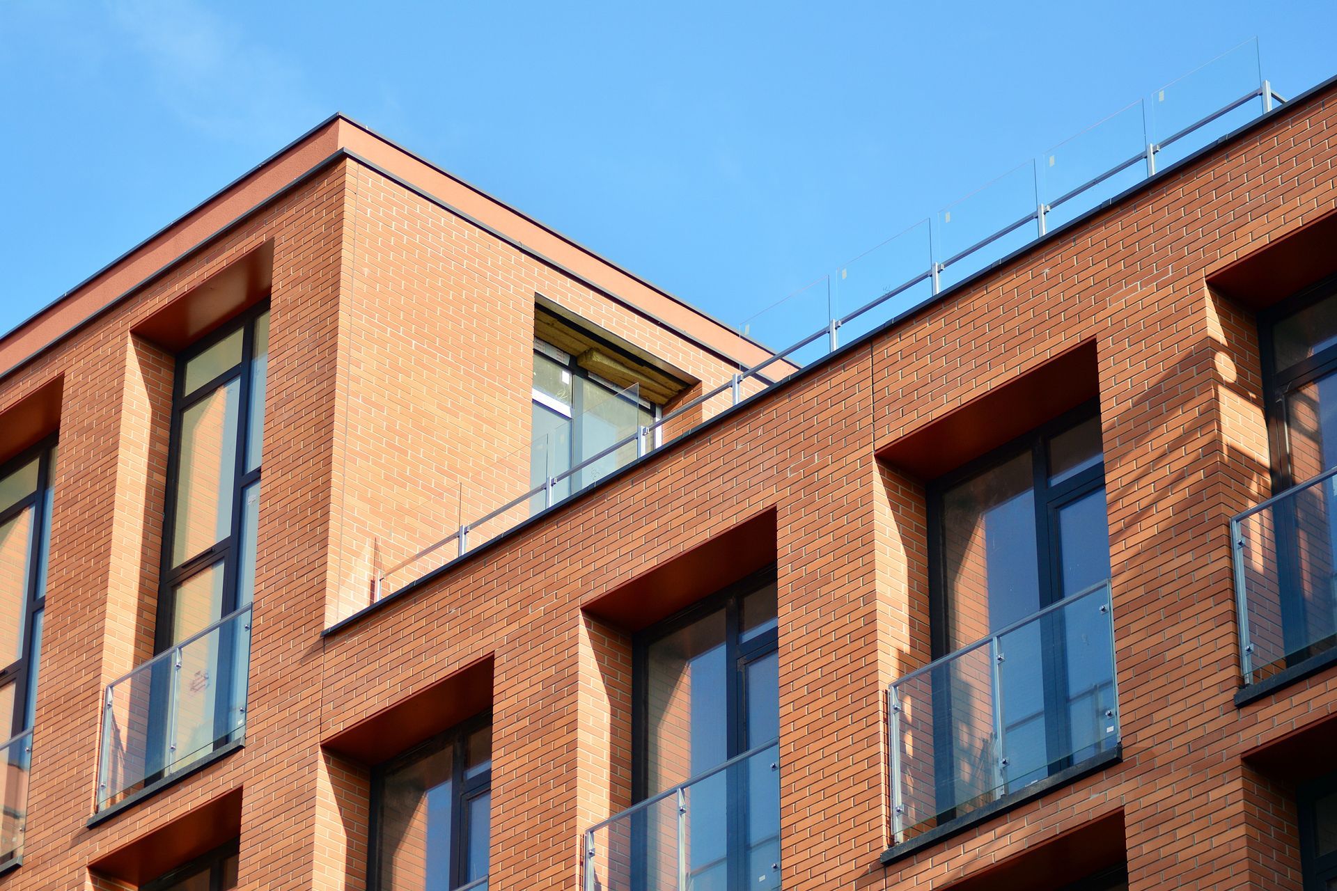 Façade en briques d'un bâtiment avec balcons et fenêtres à balustrades de verre, se détachant sur un ciel bleu.