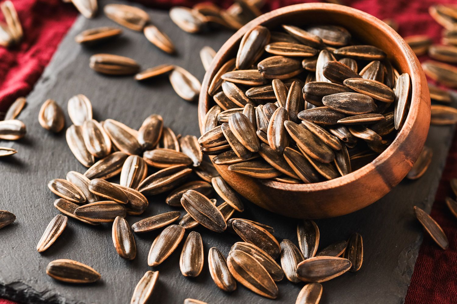 A wooden bowl filled with sunflower seeds, surrounded by scattered seeds on a dark stone surface with a red cloth backdrop.