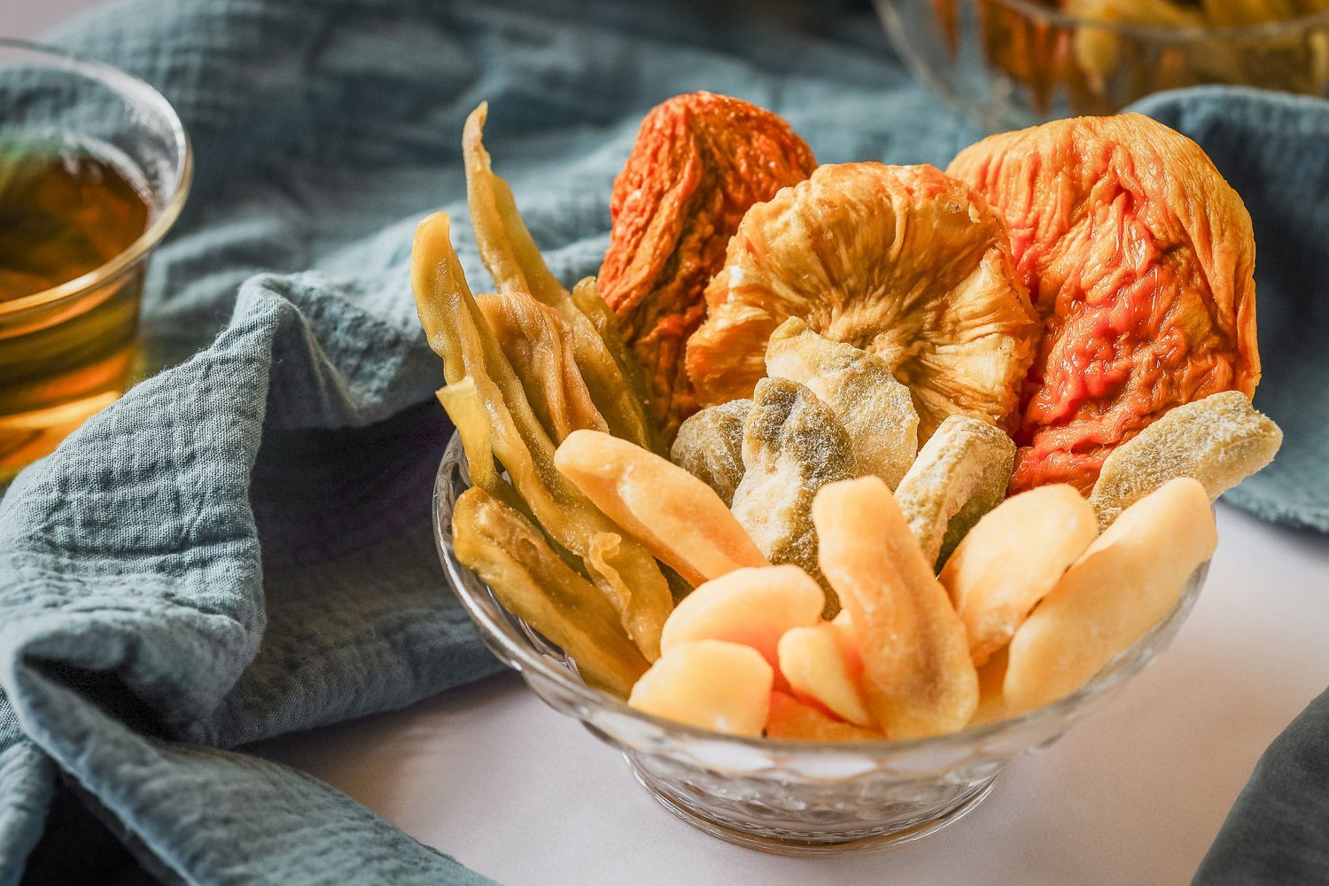 A crystal bowl filled with various dried fruits sits on a blue cloth near a small glass of tea.
