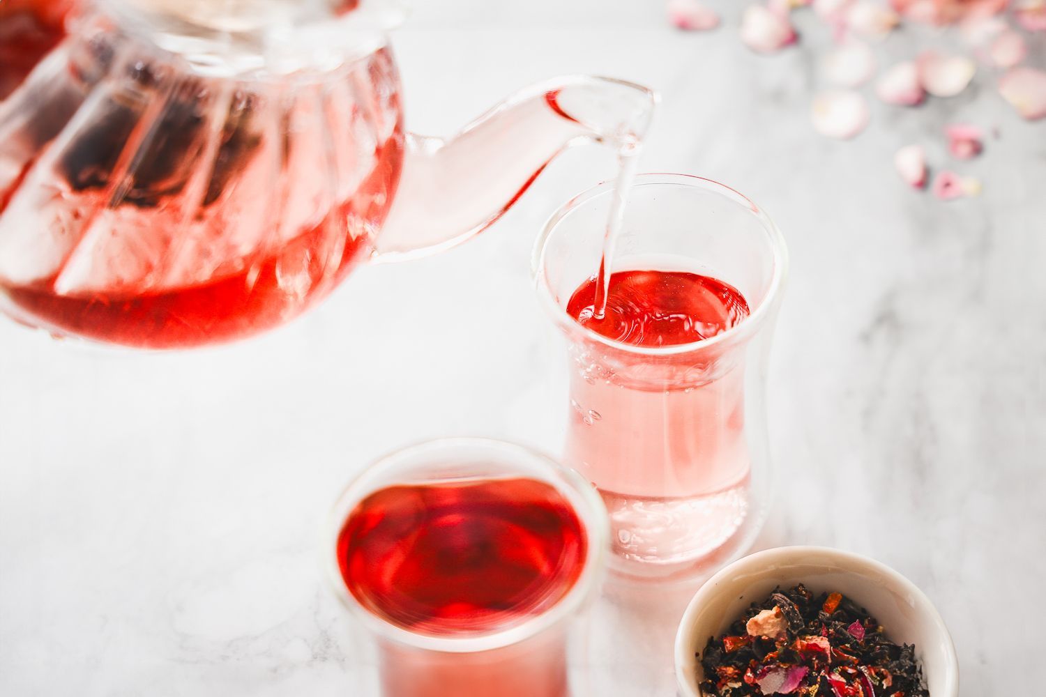 A glass teapot pours bright pink hibiscus tea into a clear glass, with another filled glass and loose tea leaves nearby.