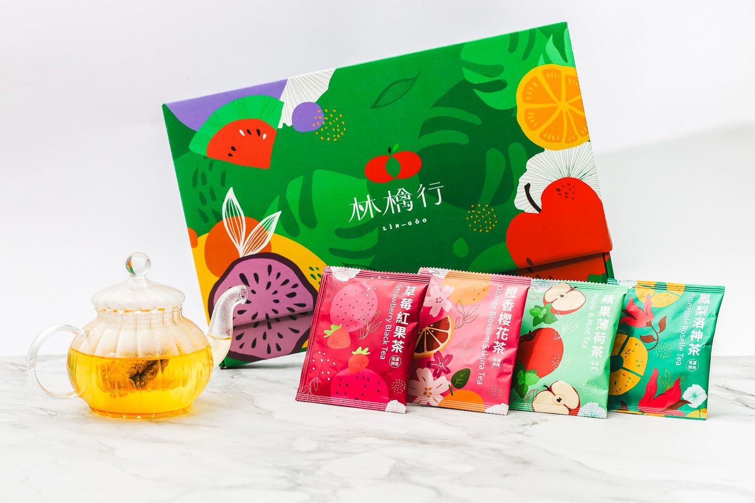 A colorful box of fruit-flavored tea bags and a glass teapot filled with amber tea on a white marble surface.