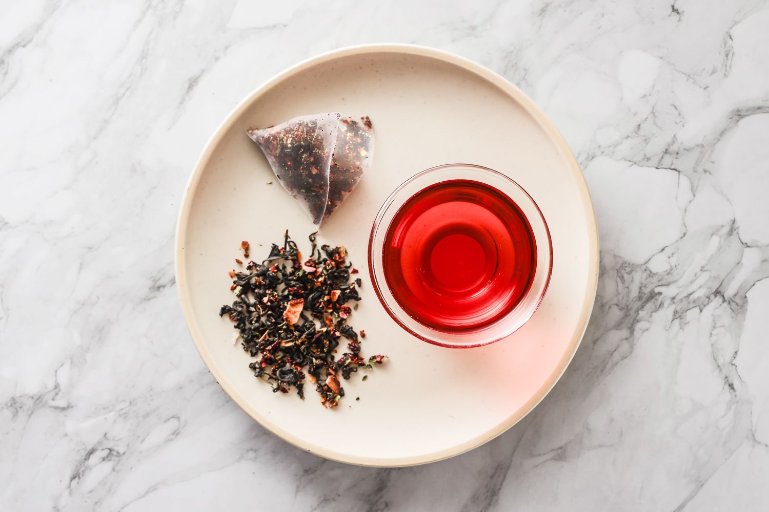 A tea bag, loose tea leaves, and a small glass of vibrant red tea arranged on a cream-colored plate on a marble surface.