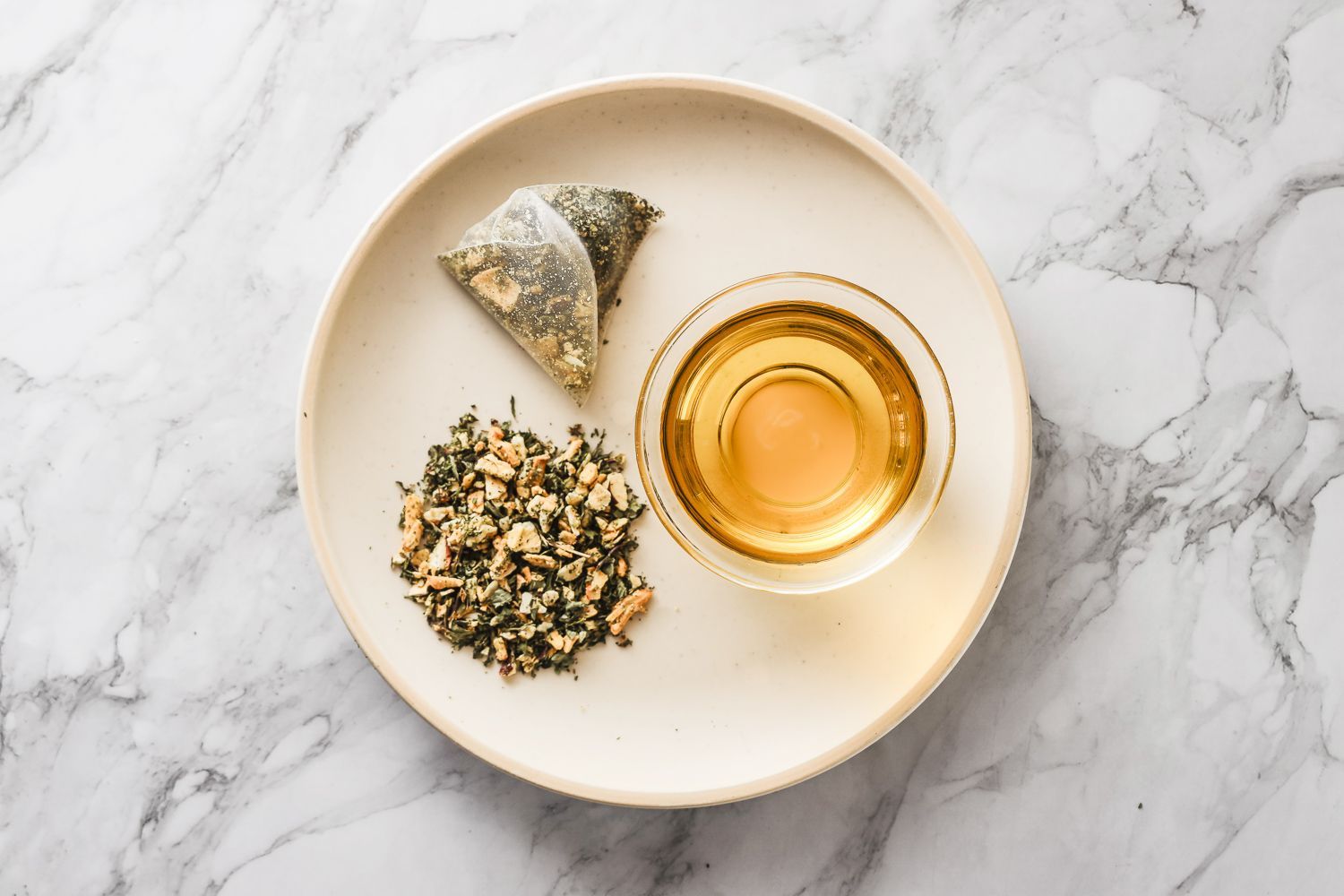 A tea bag, loose tea leaves, and a small glass of brewed tea are arranged on a plate against a white marble background.