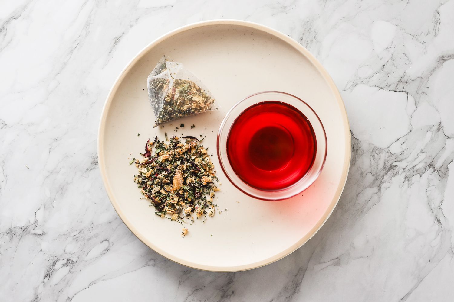 A plate holding a pyramid tea bag, a small pile of loose-leaf herbal tea, and a glass bowl of bright red tea.
