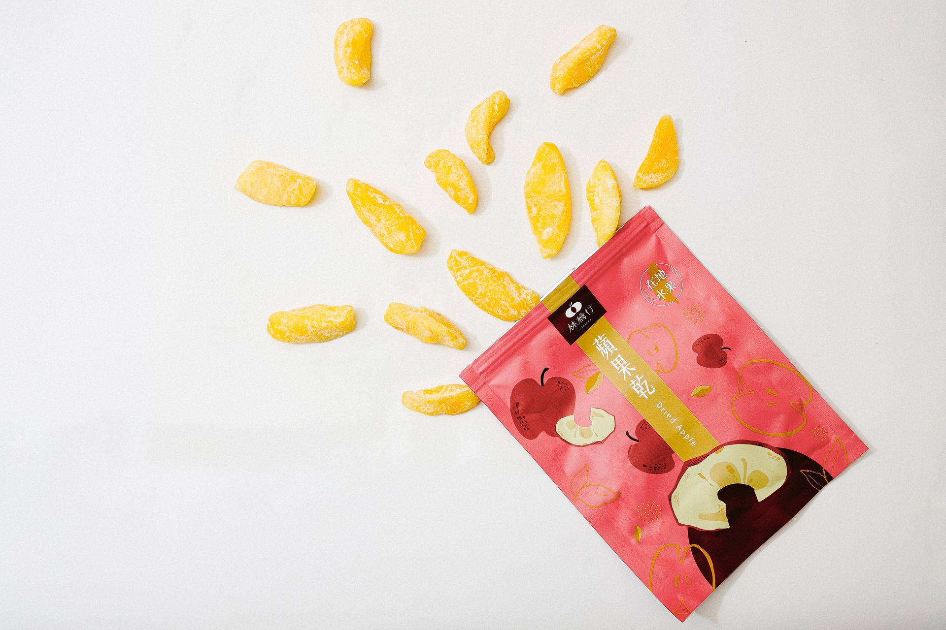 A pink snack bag for dried apple slices sits on a white surface, with scattered pieces flowing out of it.