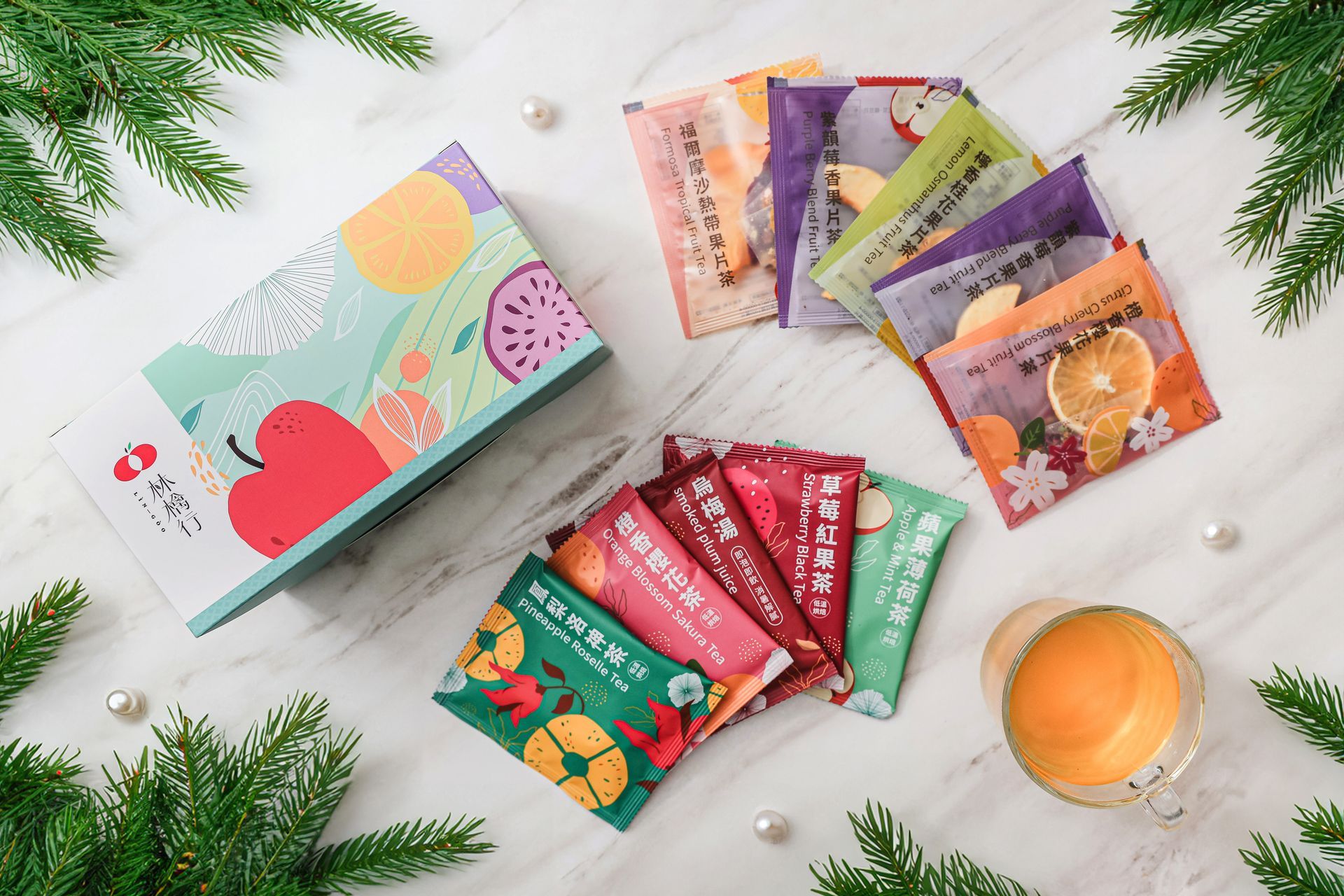 A gift box and various colorful tea sachets arranged on a white marble surface decorated with evergreen branches.