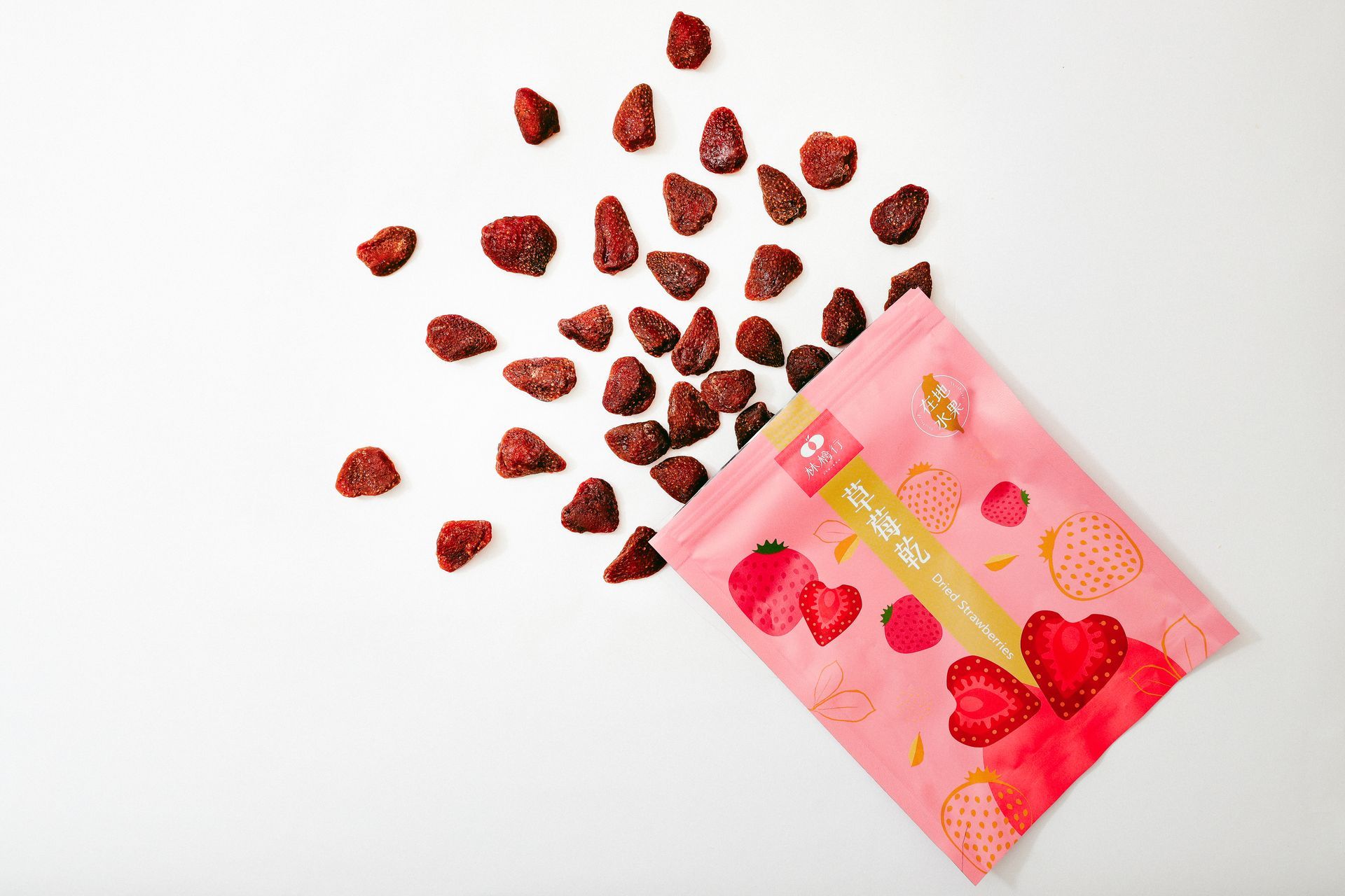 A pink bag of freeze-dried strawberries spilled onto a plain white background, showing several individual dried pieces.