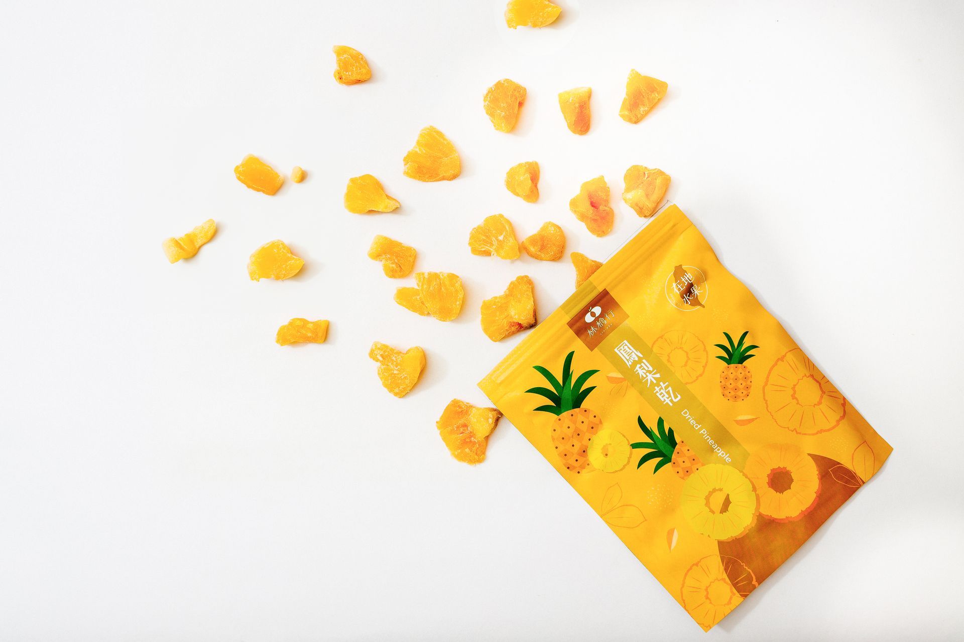 Dried pineapple chunks scattered on a white surface next to a yellow pineapple-themed snack bag.