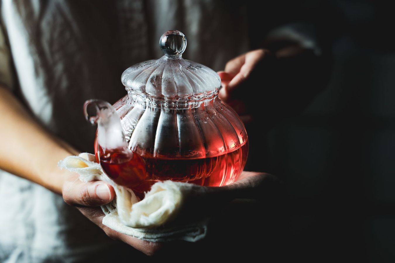 一個人手捧一個裝滿深紅色果片茶的玻璃茶壺，茶壺放在一塊柔軟的白色布料中，背景是深色的。